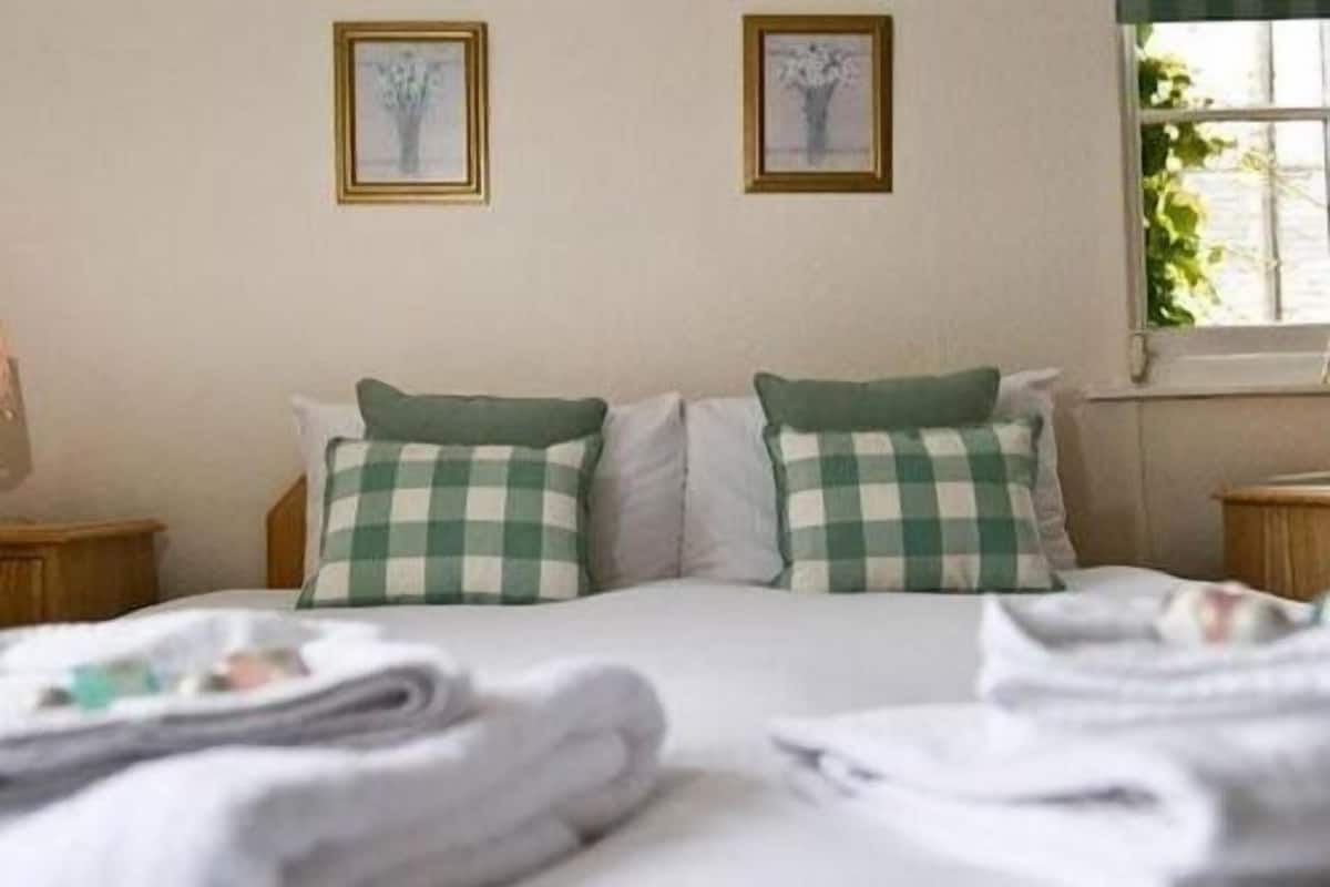 Ein ordentlich bezogenes Bett mit karierten Kissen, ein Fenster mit Blick auf Grünflächen und zwei gerahmte Bilder an der Wand.