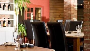 A restaurant interior with brick walls, wooden tables, and black chairs. There is a shelf with bottles in the background.