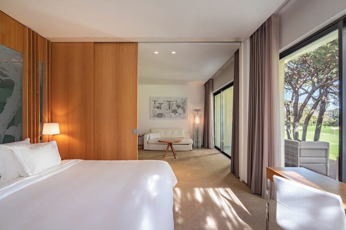 Ein Hotelzimmer mit einem großen Bett, einer Couch, einem Esstisch und einem großen Fenster mit Blick auf Bäume.