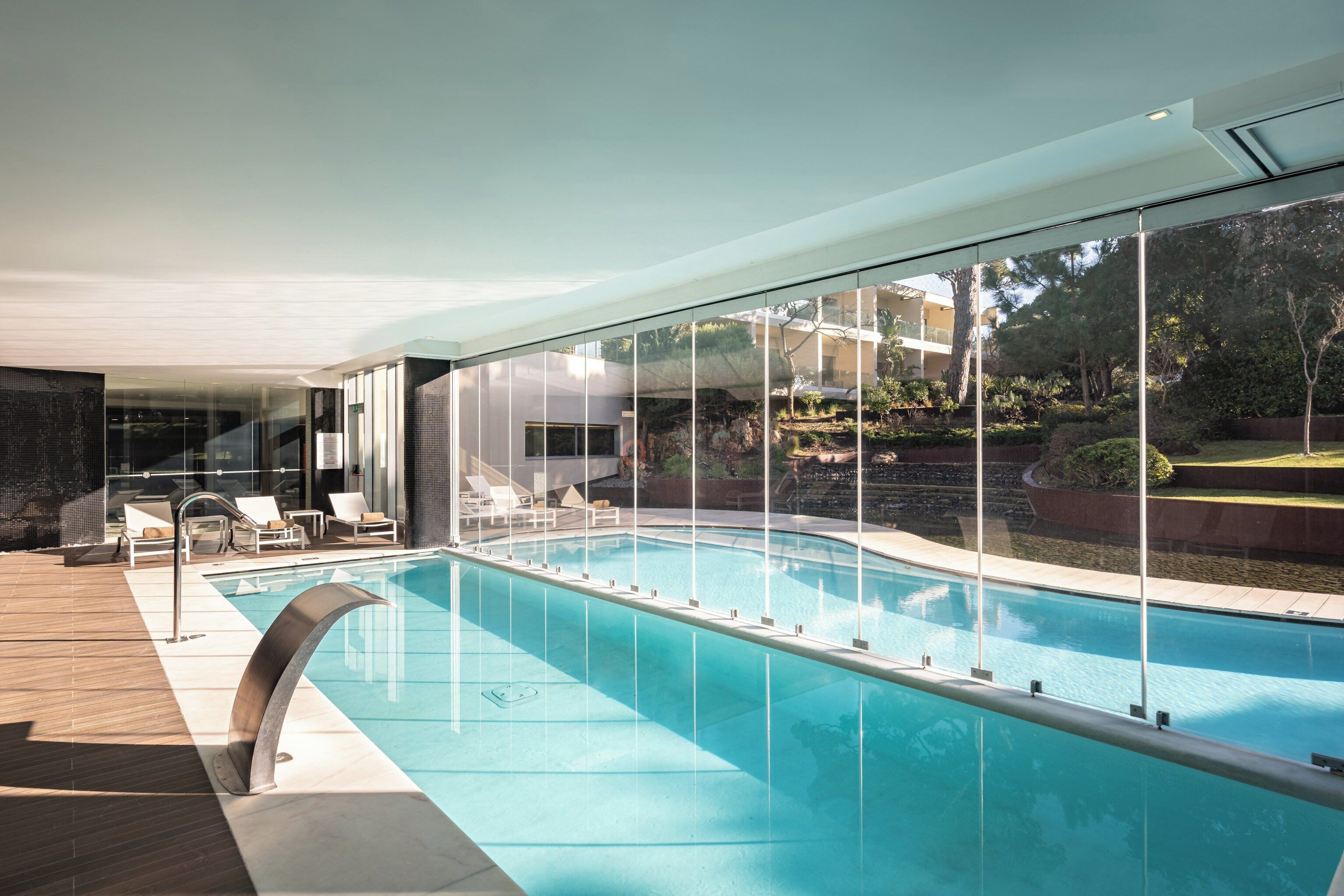 A modern indoor swimming pool with clear glass walls, lounge chairs, and a view of the surrounding landscape.