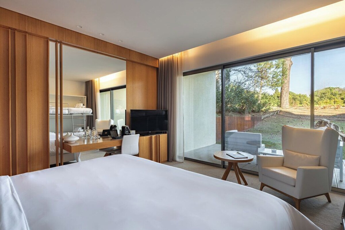 Ein Hotelzimmer mit einem Bett, einem Schreibtisch mit Fernseher, einem Sessel, einem kleinen Tisch und einem großen Fenster mit Blick ins Freie.