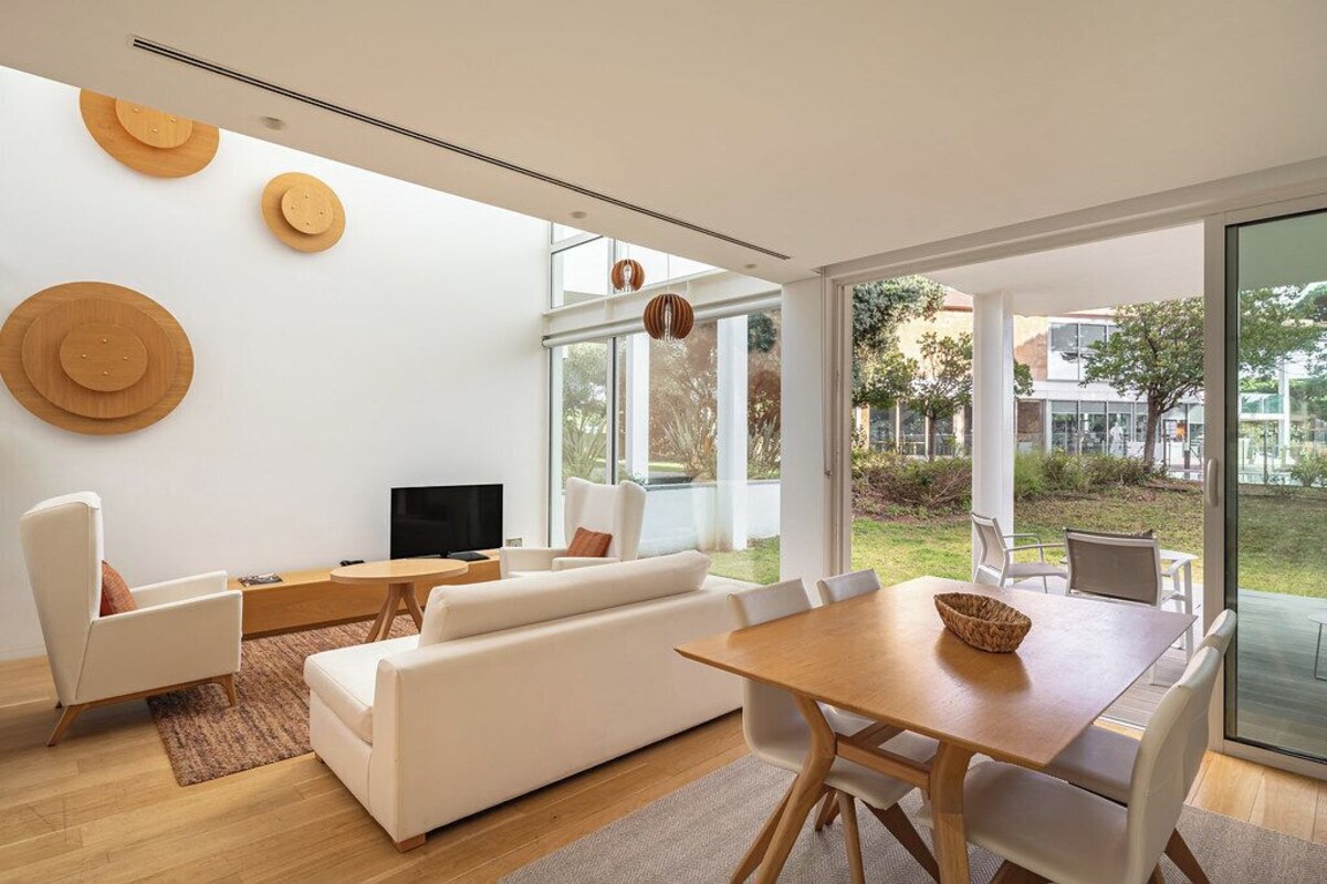 Ein modernes Wohnzimmer mit Sofa, Esstisch, Stühlen, einem Flachbildfernseher und großen Fenstern mit Blick ins Freie.