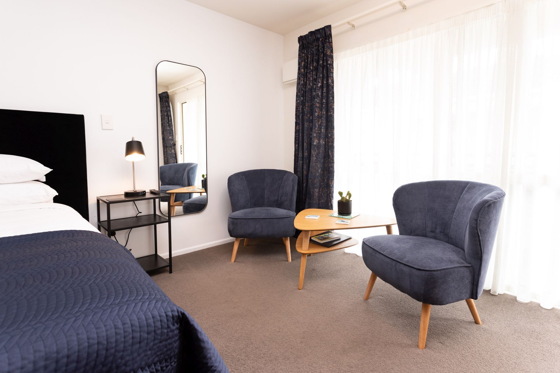 Habitación de hotel con cama, dos sillones, una mesita, lámpara, espejo y ventana con cortinas.