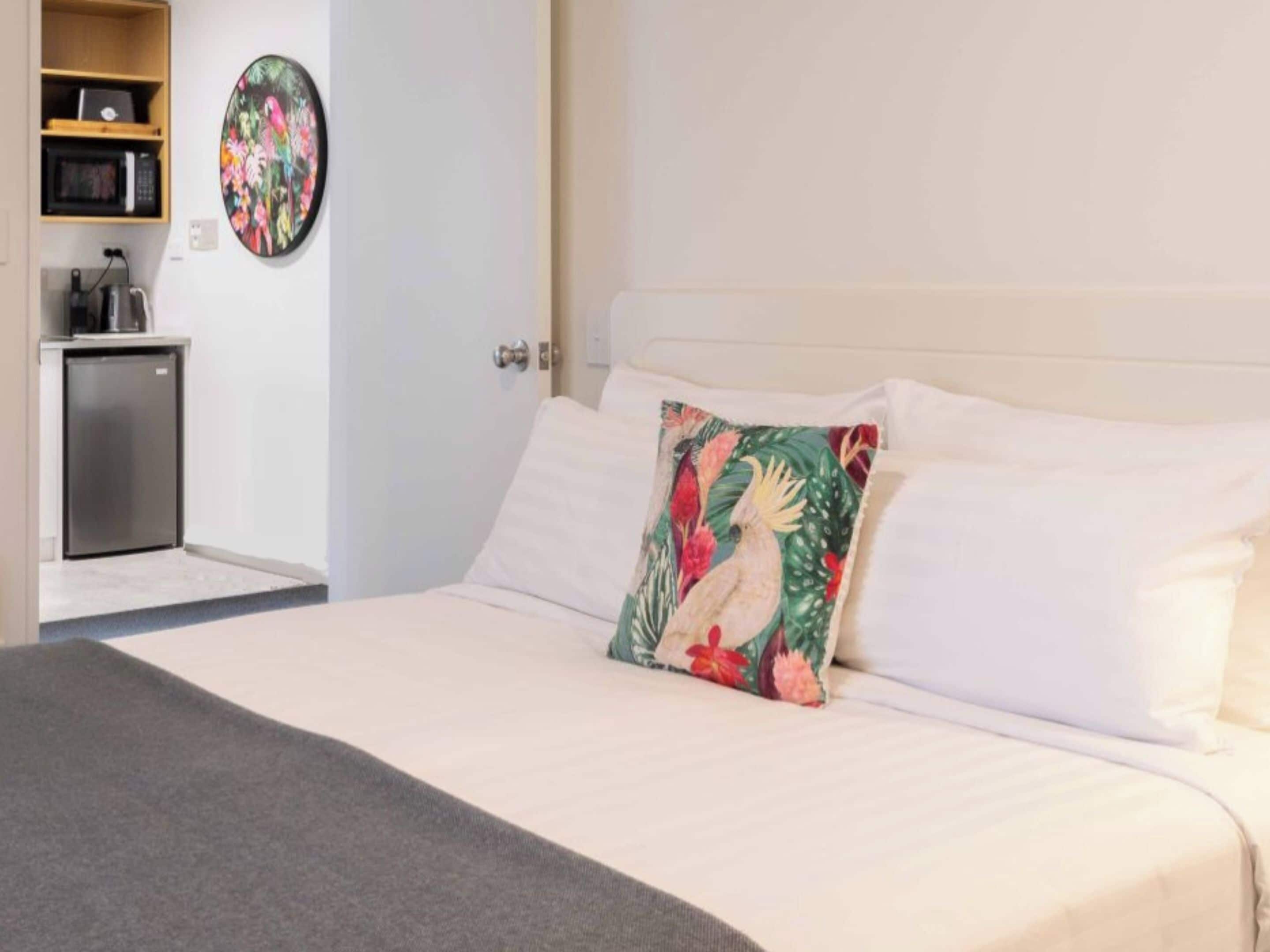 Habitación de hotel con cama, almohada con diseño tropical, microondas, refrigerador y una pequeña zona de kitchenette.