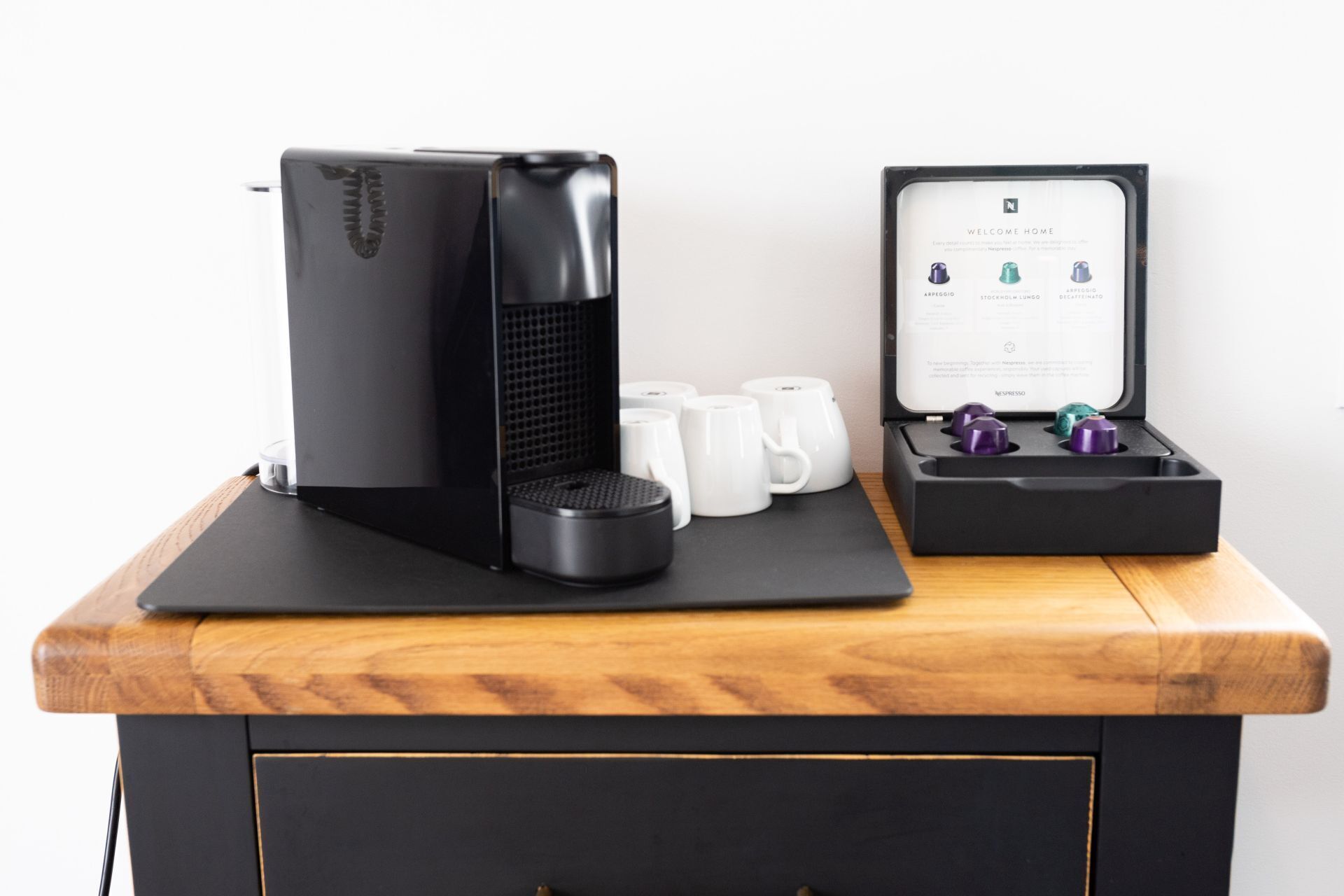 A coffee machine on a wooden table with a black tray, a box with coffee capsules, and white mugs.