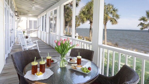 Un espacio de comedor al aire libre cubierto con vistas al mar, con sillas de mimbre, una mesa de cristal y un jarrón de tulipanes rosados.