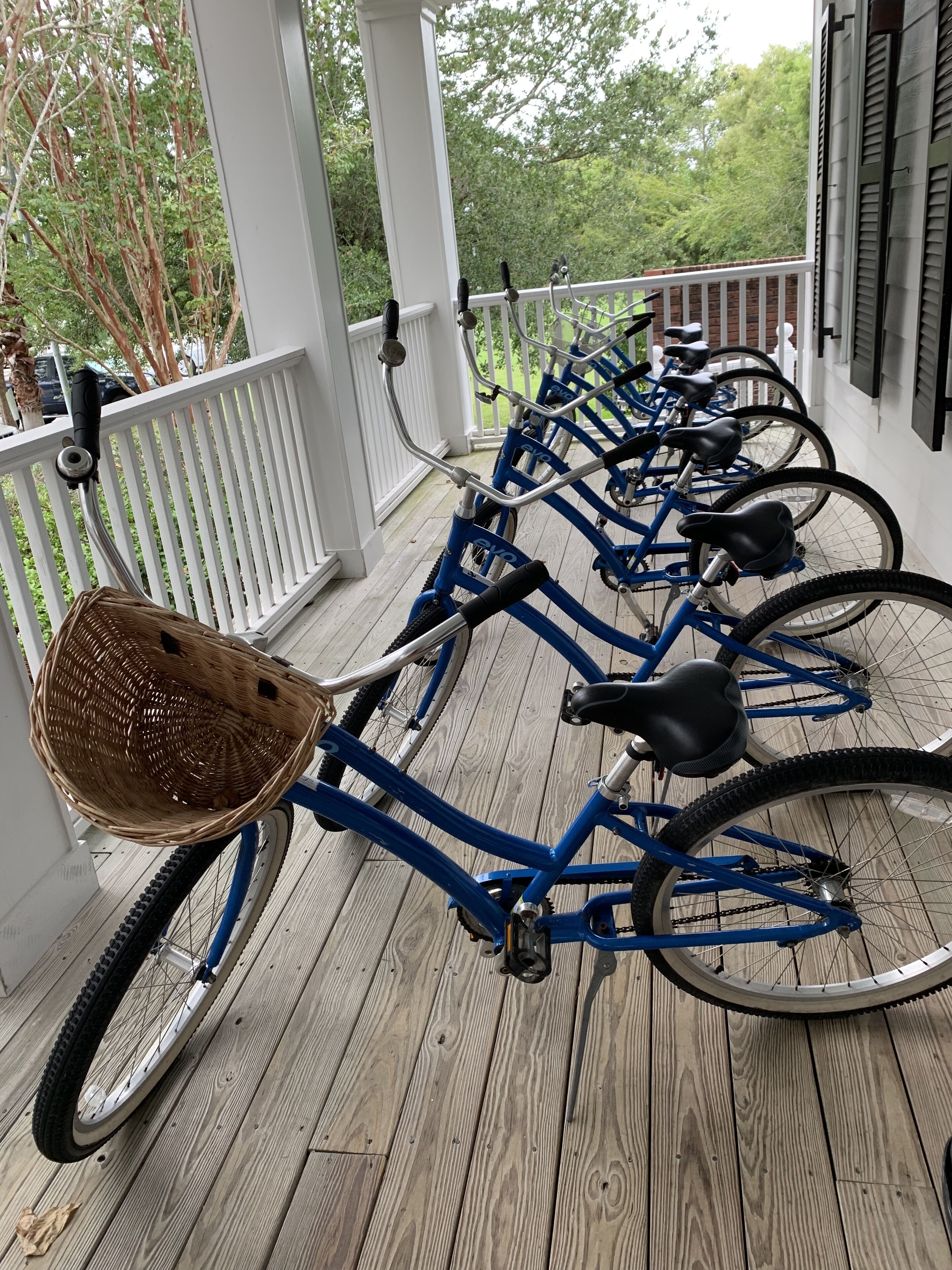 Una hilera de bicicletas azules estacionadas en un muelle de madera con barandillas blancas, una canasta de mimbre en una bicicleta y un muro de ladrillo al fondo.