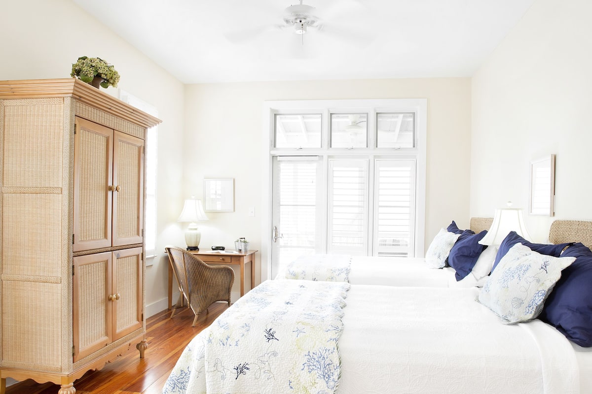 Quarto com piso de madeira, cama com colcha floral branca e azul, guarda-roupa de madeira, escrivaninha com luminária e janela com persianas.