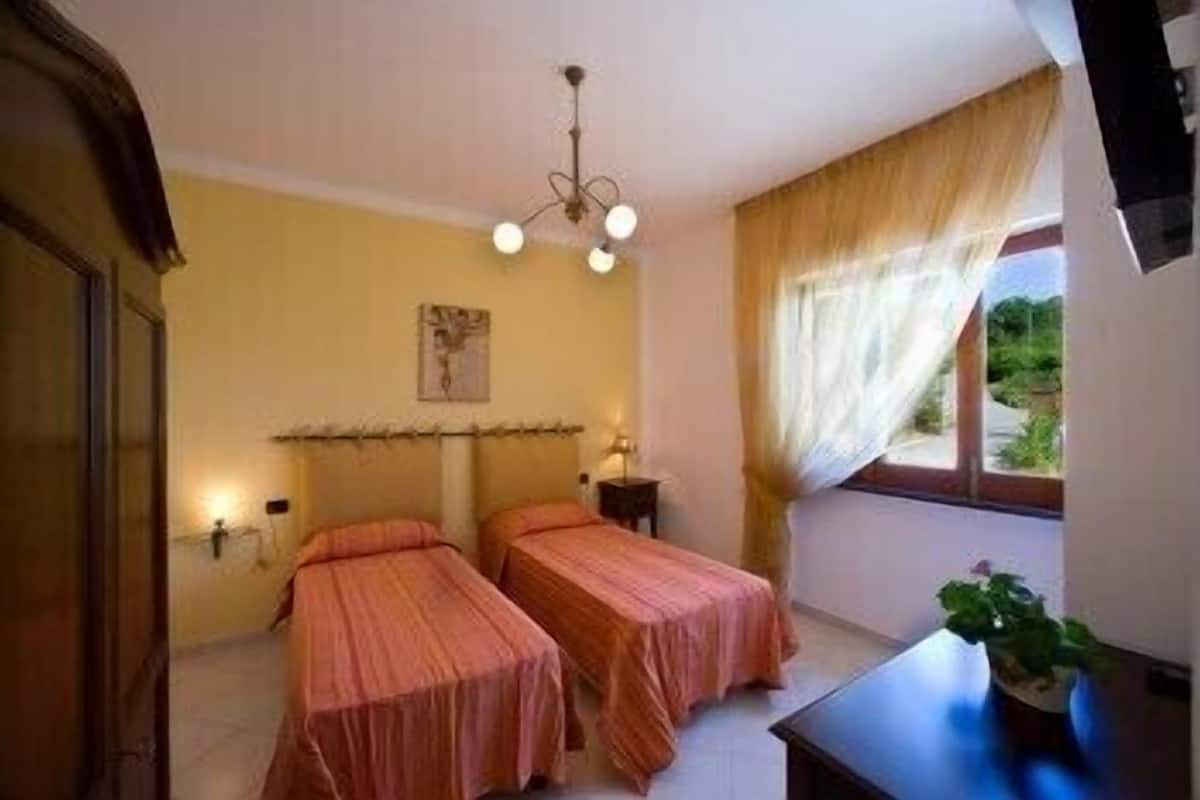 A room with two beds, a small table, a window with curtains, and a wall-mounted light fixture.