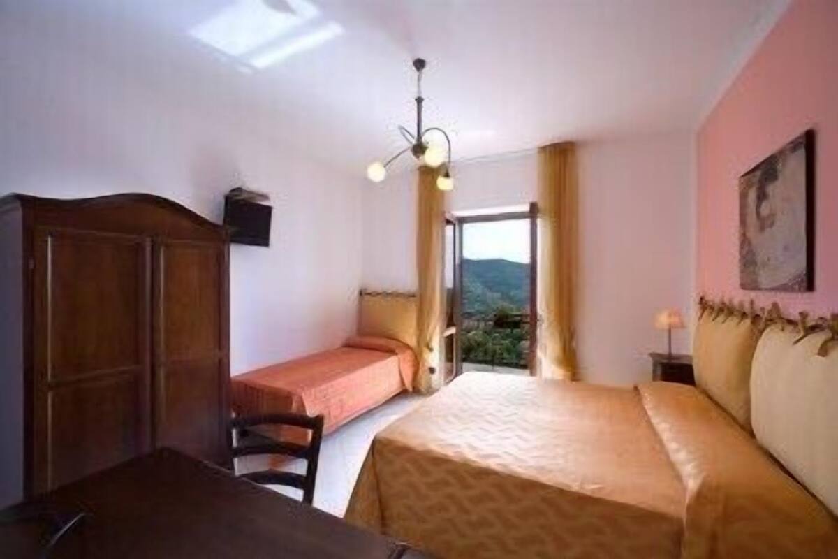 A bedroom with a bed, a desk, and a wardrobe. There is a window with a view of the outdoors.