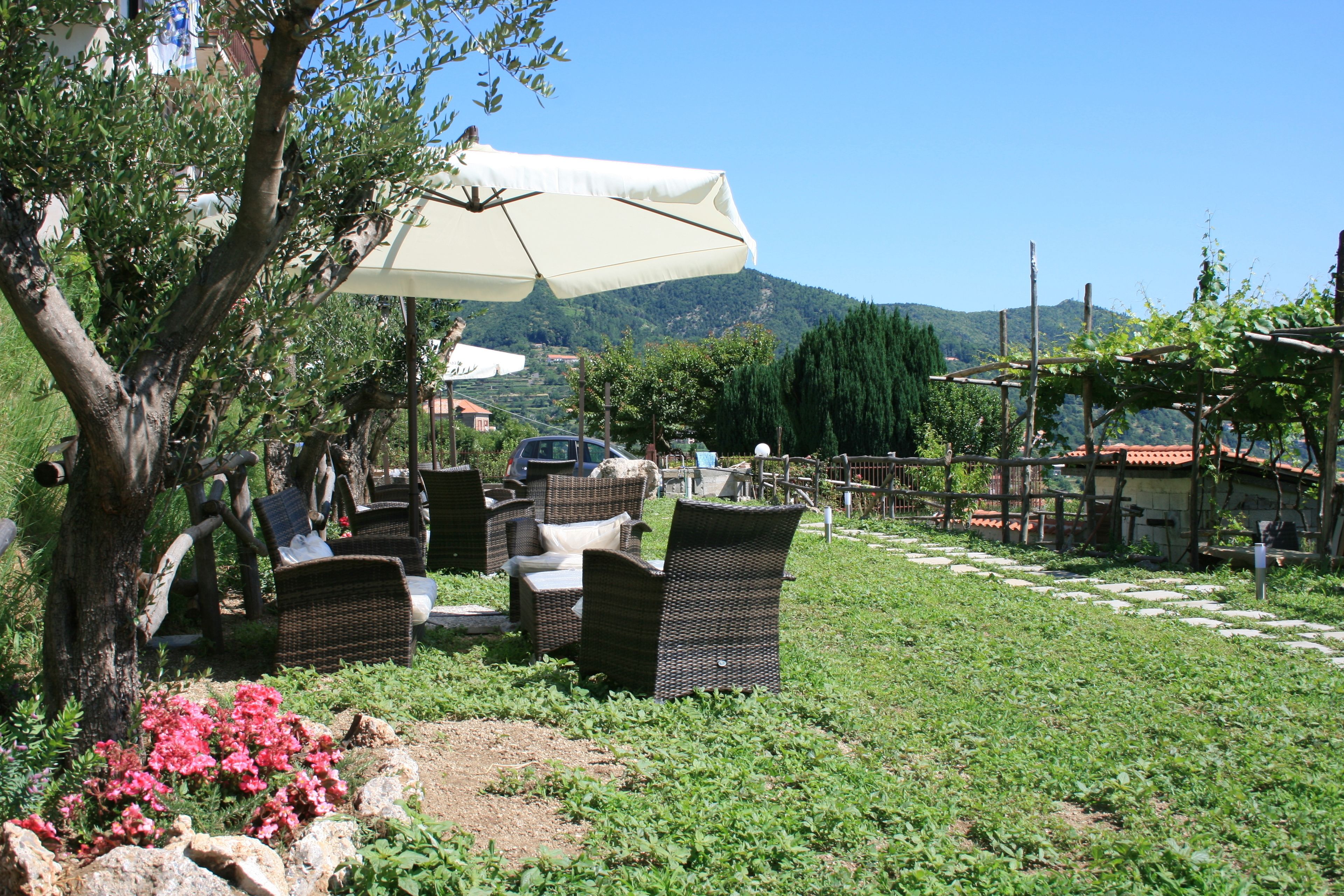 Ein Sitzbereich im Freien mit Korbmöbeln und einem großen Sonnenschirm, umgeben von viel Grün und mit Blick auf Berge.