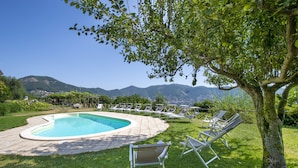 A swimming pool with lounge chairs and a tree in the foreground, surrounded by a grassy area and mountains in the background. - Casa Pendola (Agerola)