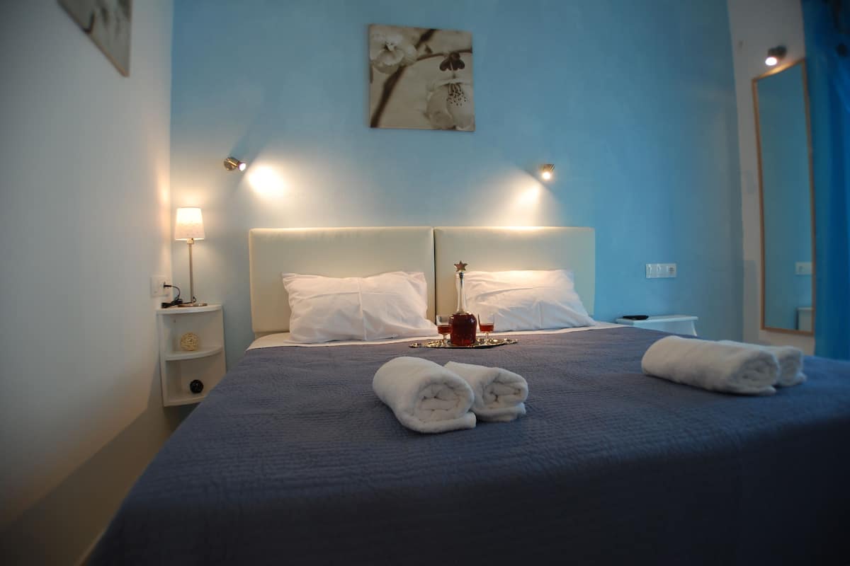 A neatly made bed with white linens and towels, a bedside table with a lamp, and a wall-mounted artwork.