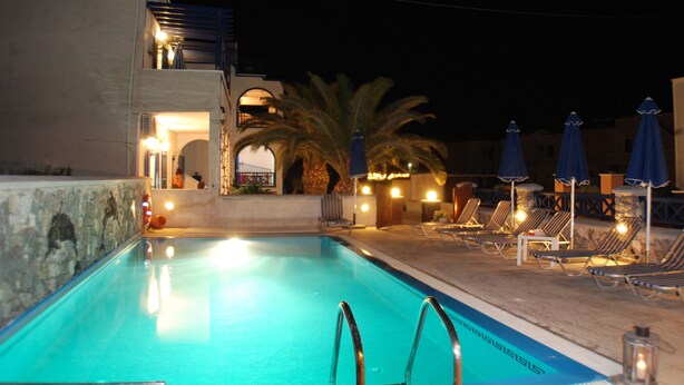Una piscina con sillas de descanso y sombrillas, un muro de piedra y un edificio iluminado.