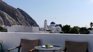 Una terraza con dos sillas de madera y una mesita, con vistas a un pueblo costero de edificios blancos y cúpulas azules, y una montaña al fondo.