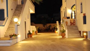 Vista nocturna de una calle estrecha con escalones que conducen a un edificio. Hay plantas en macetas y muebles de exterior a lo largo de los lados.