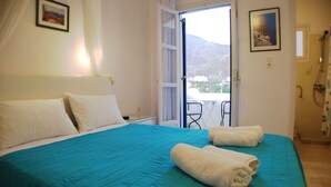 Habitación de hotel con una cama, dos almohadas, una colcha color verde azulado, dos toallas enrolladas, una ventana con vistas a montañas y una puerta que da a un balcón.