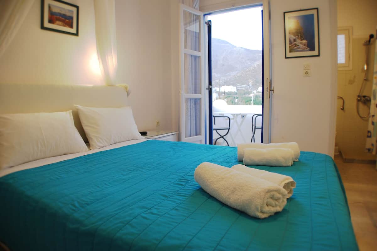 A hotel room with a bed, two pillows, a teal bedspread, two rolled towels, a window with a view of mountains, and a door leading to a balcony.