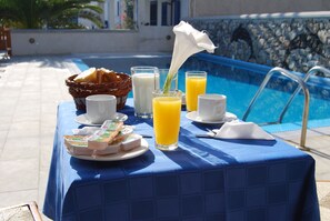 Desayuno junto a una piscina, con una mesa cubierta por un mantel azul, platos de comida, vasos de leche y zumo de naranja, y una cesta de pan.