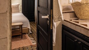 A narrow hallway leading to a bedroom with a bed, a bedside table, a lamp, and a woven basket.