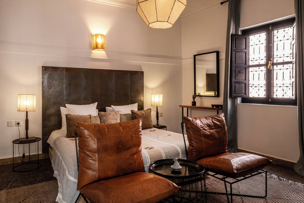 Habitació d'hotel amb un llit gran, dues taules de nit, una cadira, una taula petita, una làmpada, un mirall i una finestra amb baranes de ferro decoratives.
