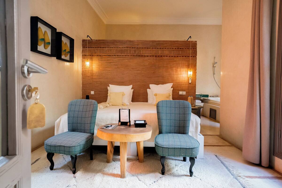 Una habitació d'hotel amb un panell de fusta al capçal del llit, dues cadires iguals, una taula rodona i un llit amb roba de llit blanca.
