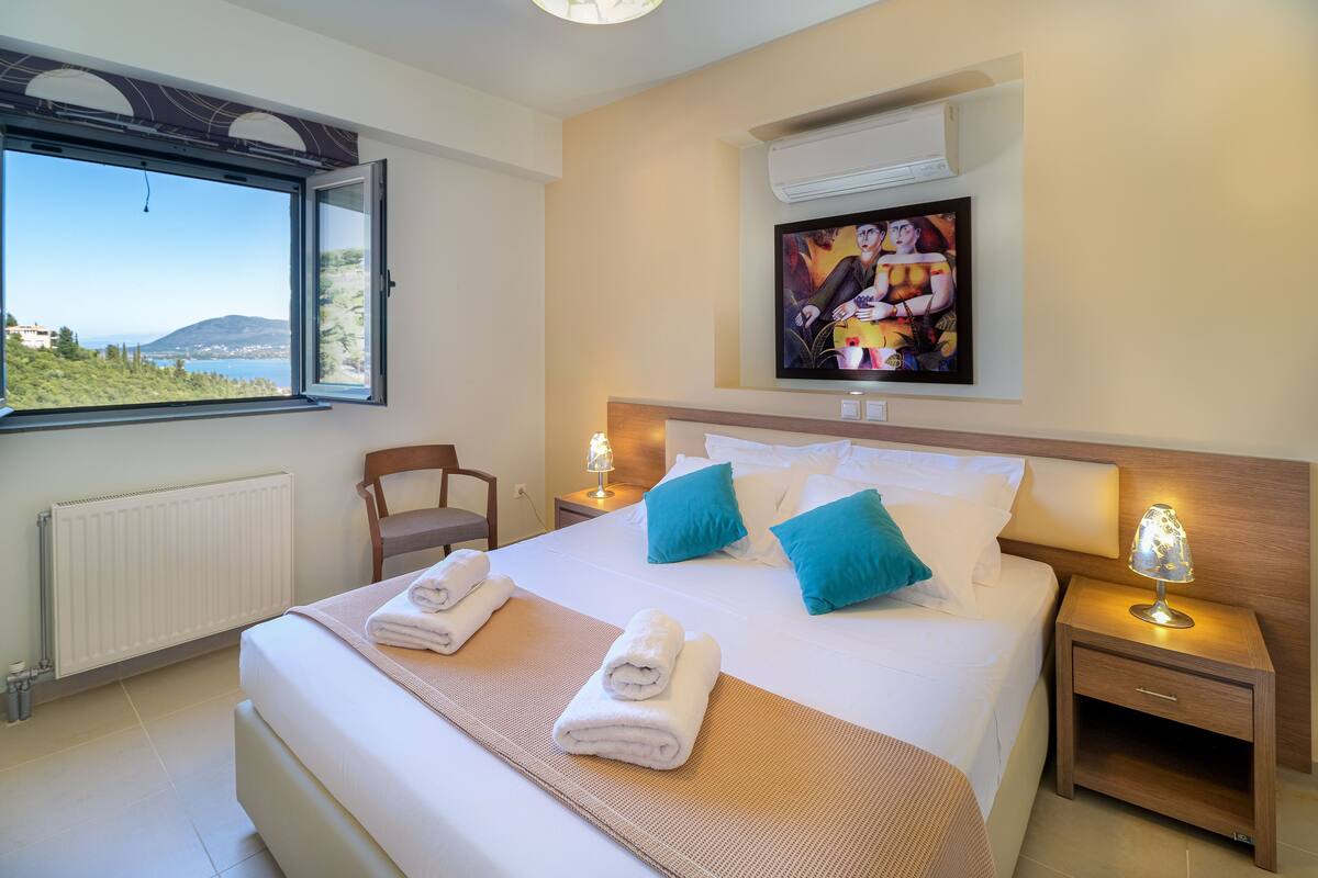 A hotel room with a bed, two bedside tables, a nightstand, a wall-mounted TV, and a window with a view of the sea and hills.