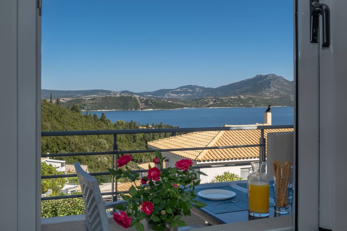 A balcony with a view of a coastal landscape, a table with a glass of orange juice, and a vase with pink flowers.