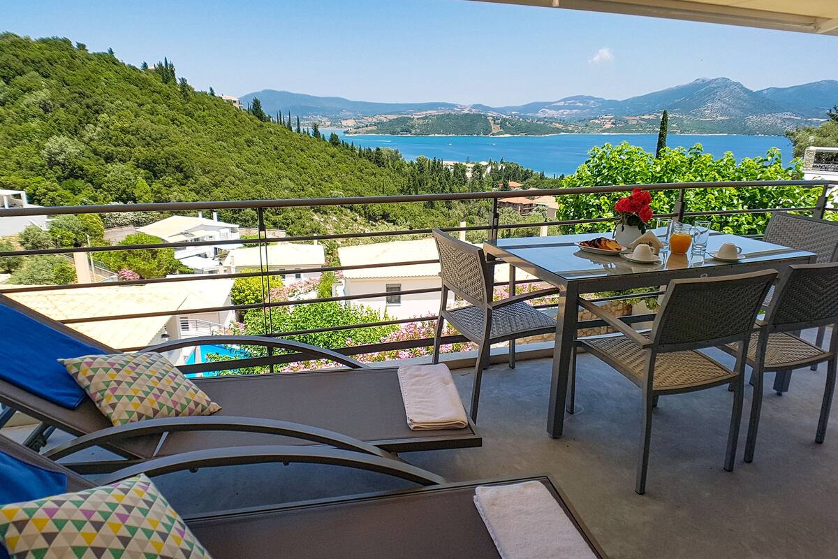 A balcony with a table set for breakfast, overlooking a scenic view of a lake and mountains.