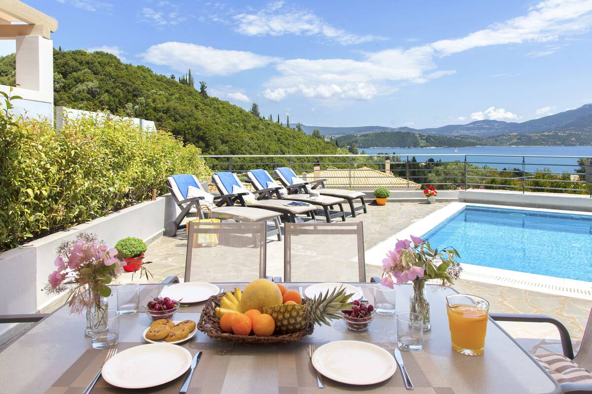A dining table set with plates, cutlery, a fruit basket, and a glass of orange juice, overlooking a pool and lounge chairs.