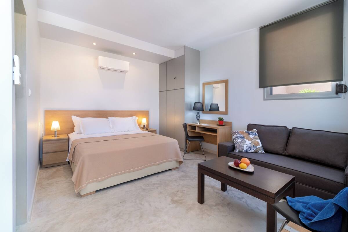 A hotel room with a bed, desk, and sofa. There is a window with a blind, a lamp, and a small table with fruit.