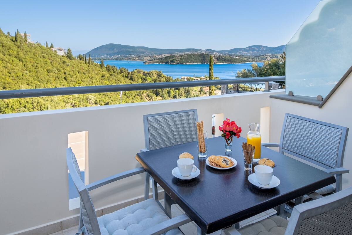 A balcony with a table set for breakfast, overlooking a scenic view of the sea and hills.