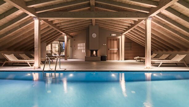 Une piscine intérieure avec des poutres en bois, des chaises longues et une cheminée.
