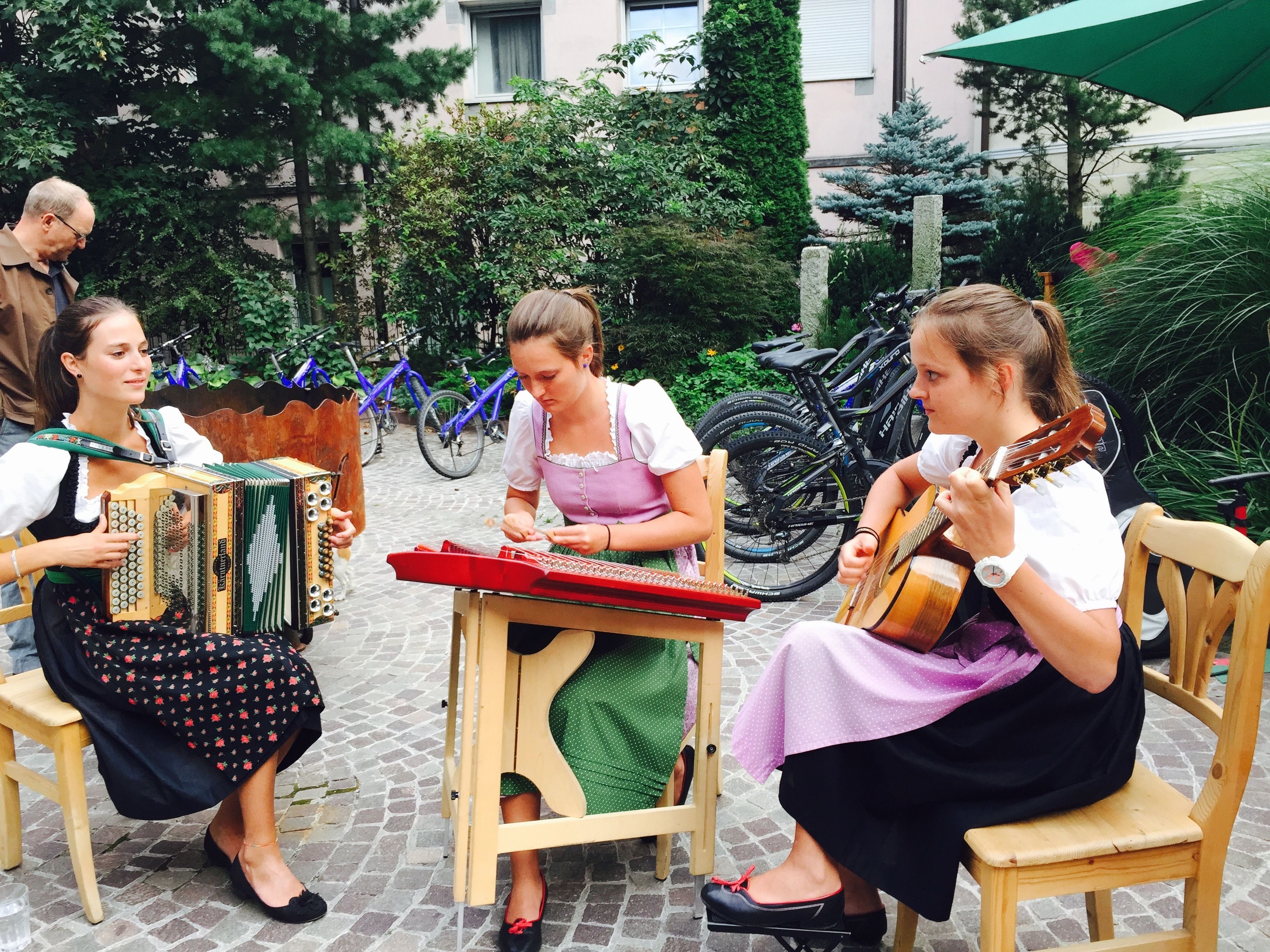 A group of people playing traditional instruments in an outdoor setting with bicycles and greenery in the background.