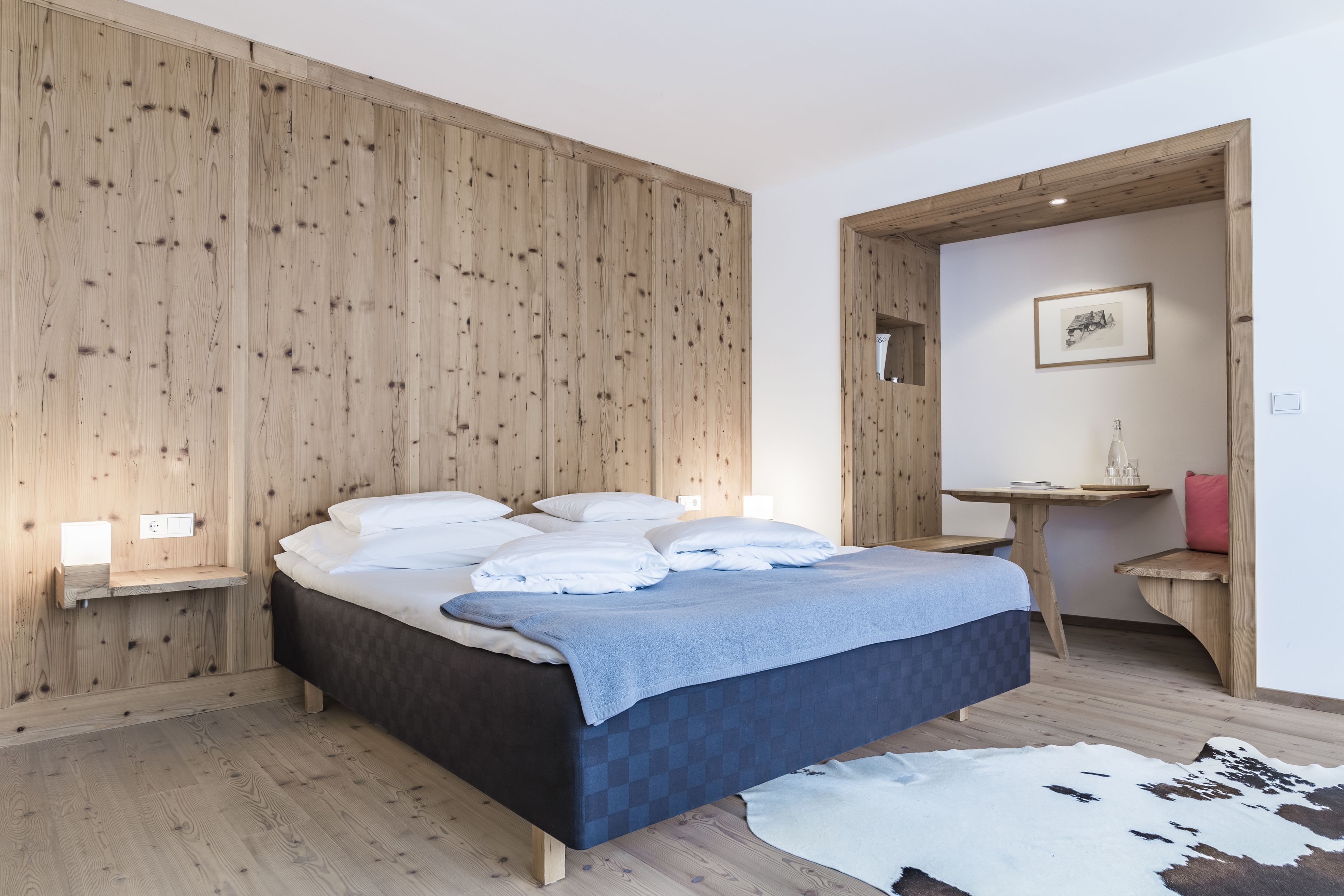 Un dormitorio con pared de madera, cama con ropa de cama blanca y azul, una mesita y un espejo.