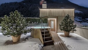 Un bain à remous en bois, accessible par des marches, entouré d’un sol enneigé et de deux arbres à feuilles persistantes plantés dans des pots.