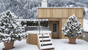 Un bâtiment en bois doté d’un bain à remous et d’un escalier menant à une porte, entouré d’arbres couverts de neige et avec une chaîne de montagnes en arrière-plan.