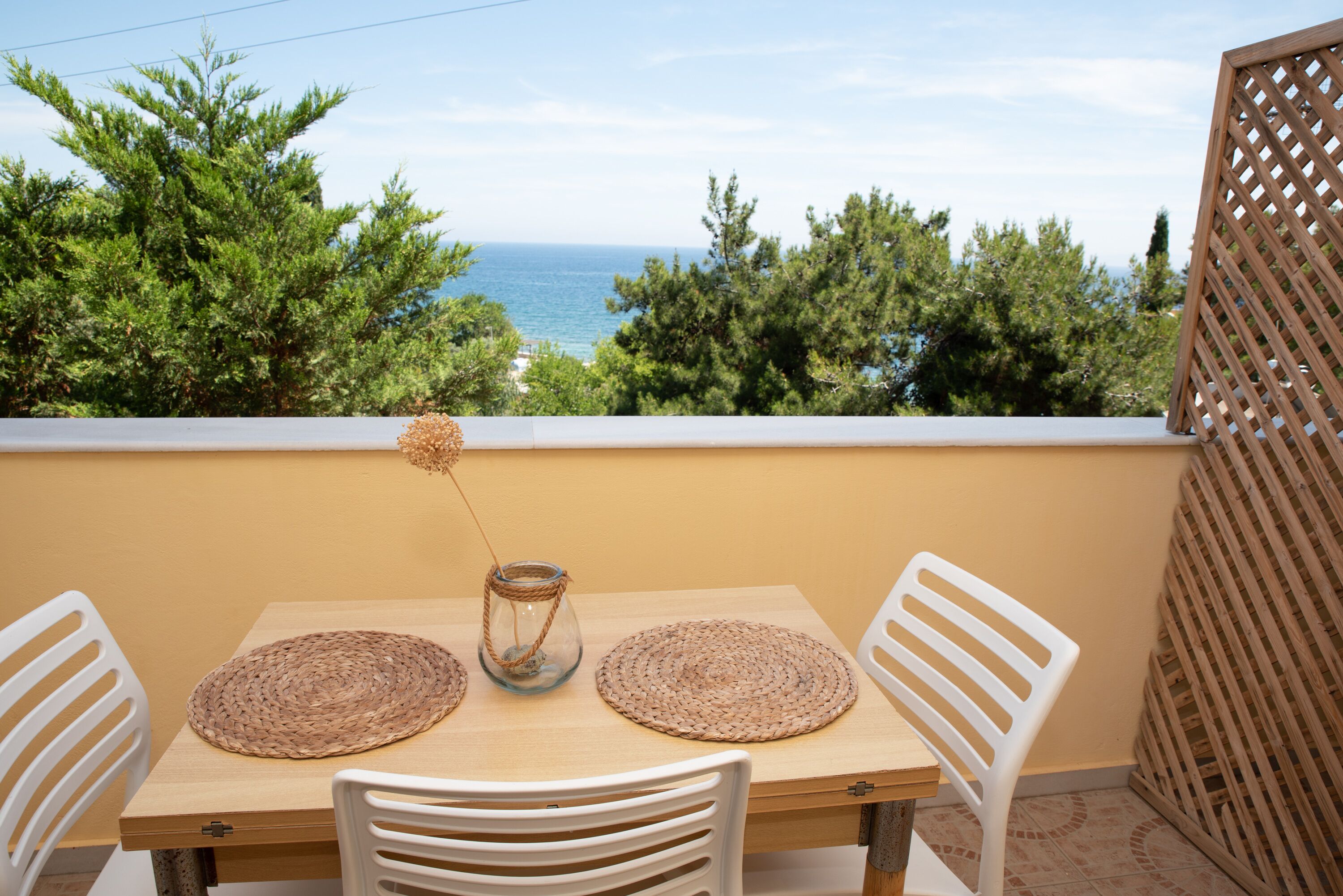 A wooden table with two woven placemats and a glass vase with a rope handle, set against a backdrop of a clear sky and a view of the sea.