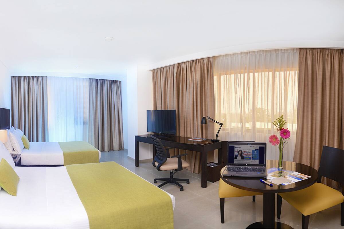 Habitación de hotel con cama, dos mesitas de noche, un escritorio con silla, una computadora portátil y un florero con flores.