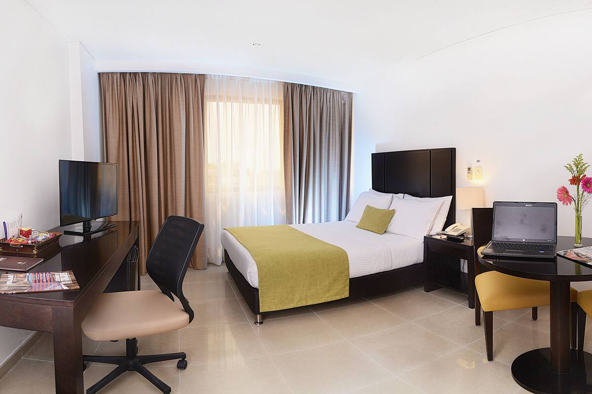 Habitación de hotel con cama, escritorio, silla, computadora portátil y una mesita con un jarrón de flores.