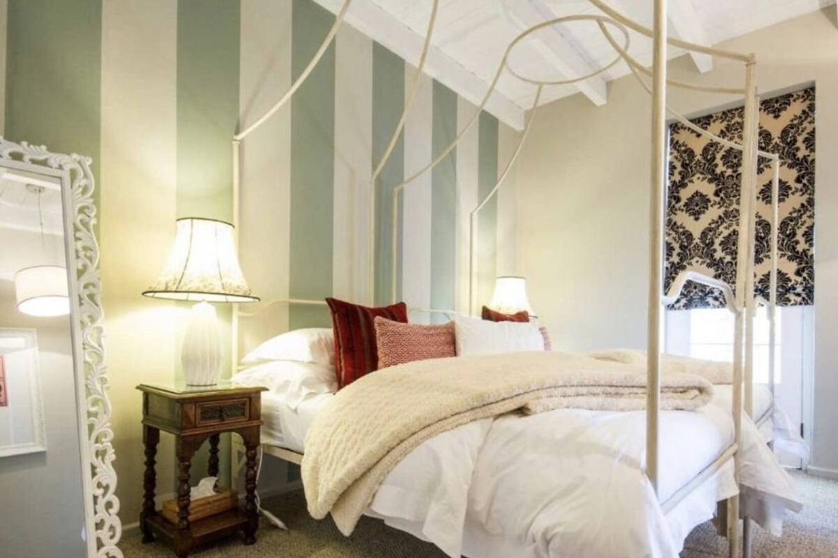 A four-poster canopy bed with white bedding, a nightstand with a lamp, and a mirror with an ornate frame.