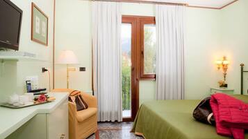 Una camera d'albergo con un letto, una scrivania con una sedia, una televisione, una finestra con tende e un comodino con una lampada.
