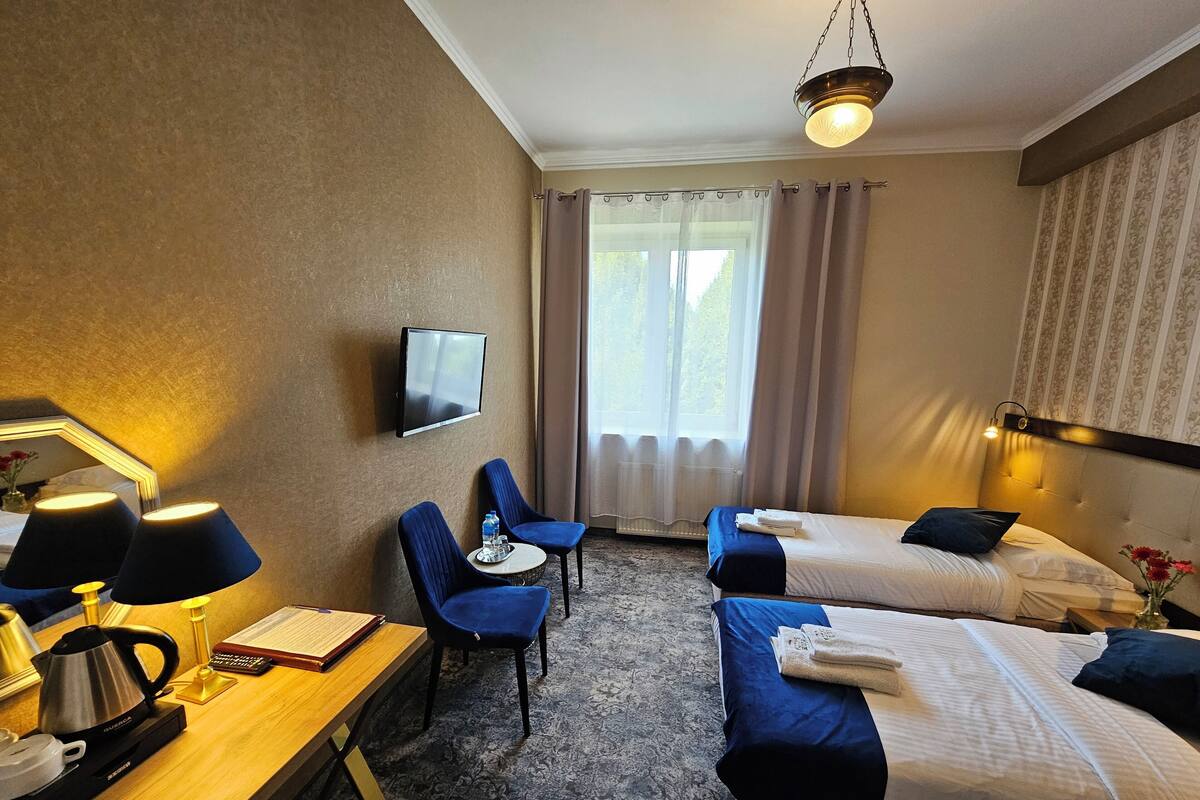 A hotel room with two beds, a desk with a kettle, a chair, a TV, and a window with curtains.