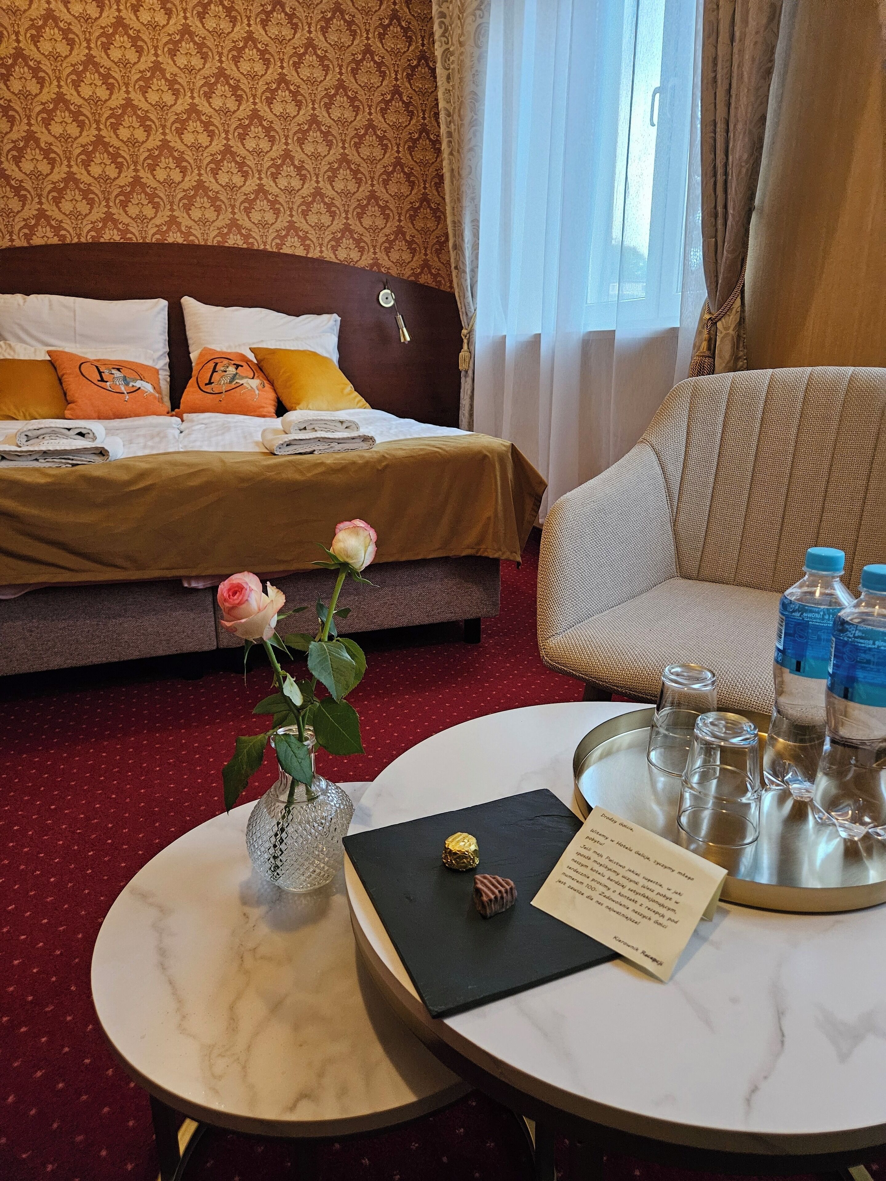 Ett hotellrum med en snyggt bäddad säng, en sittgrupp med en soffa, ett runt bord med en meny, en vas med en ros och ett bricka med vattenflaskor och glas.