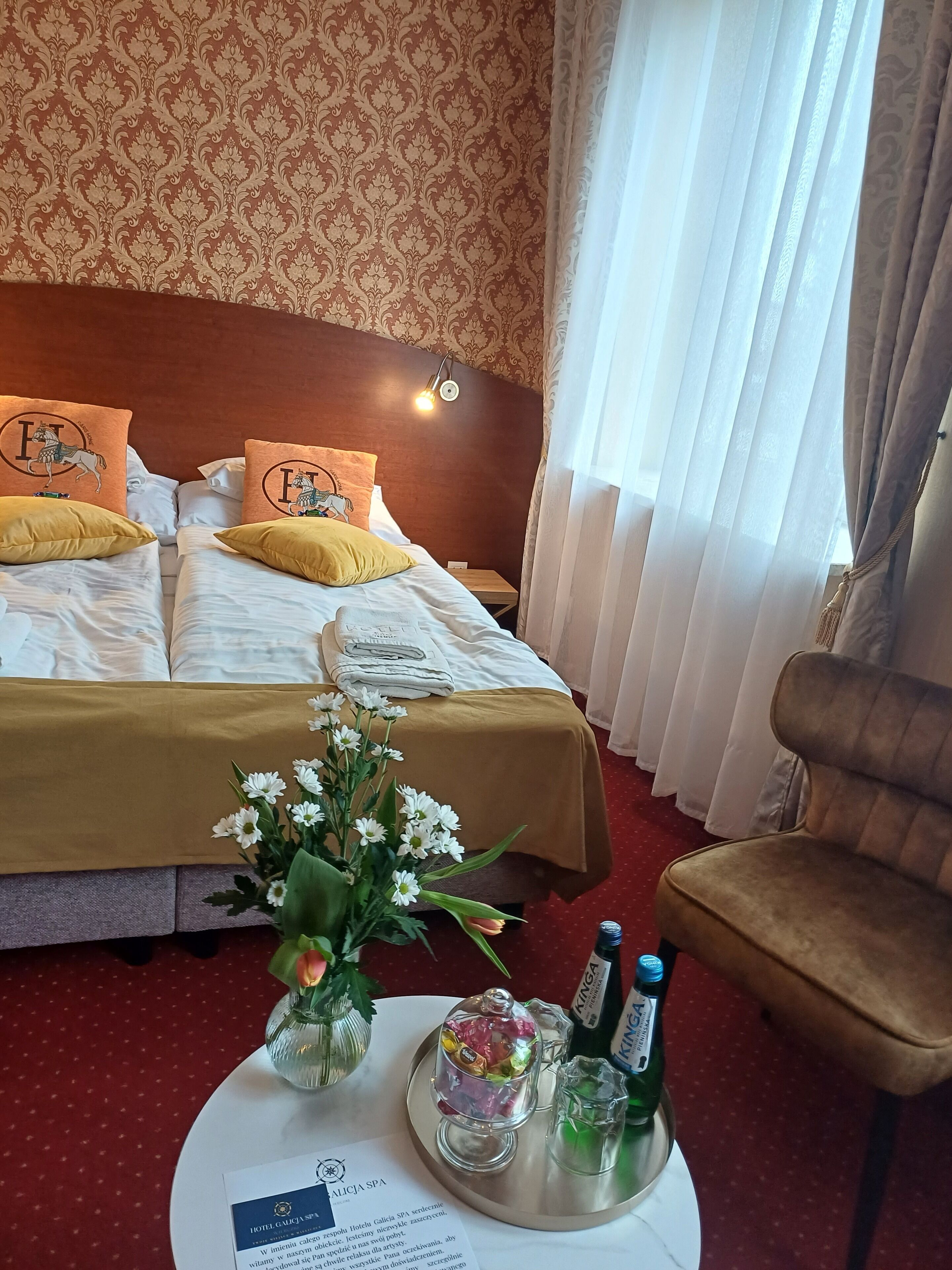 Quarto de hotel com uma cama grande, duas almofadas com desenho de cavalos, um abajur, um vaso com flores, um porta-bandeja com garrafas e uma cadeira.