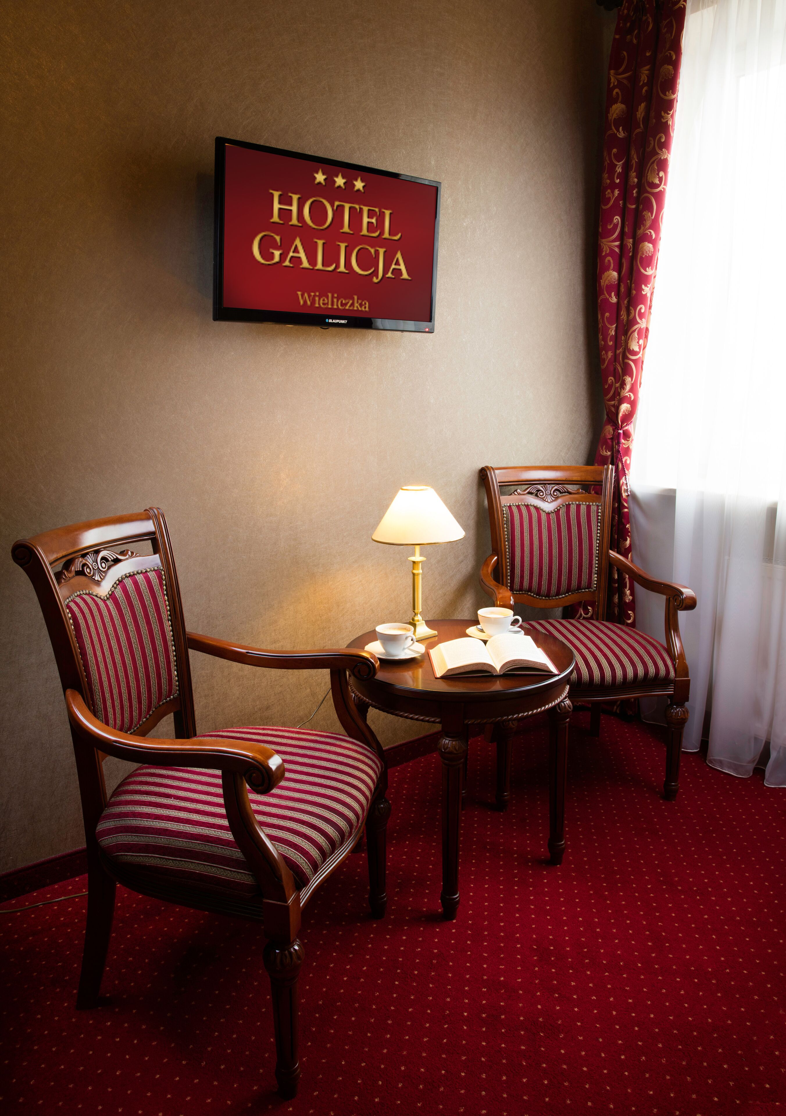A hotel room with two wooden chairs, a small table with a lamp, a book, and a sign on the wall reading "HOTEL GALICJA Wieliczka".