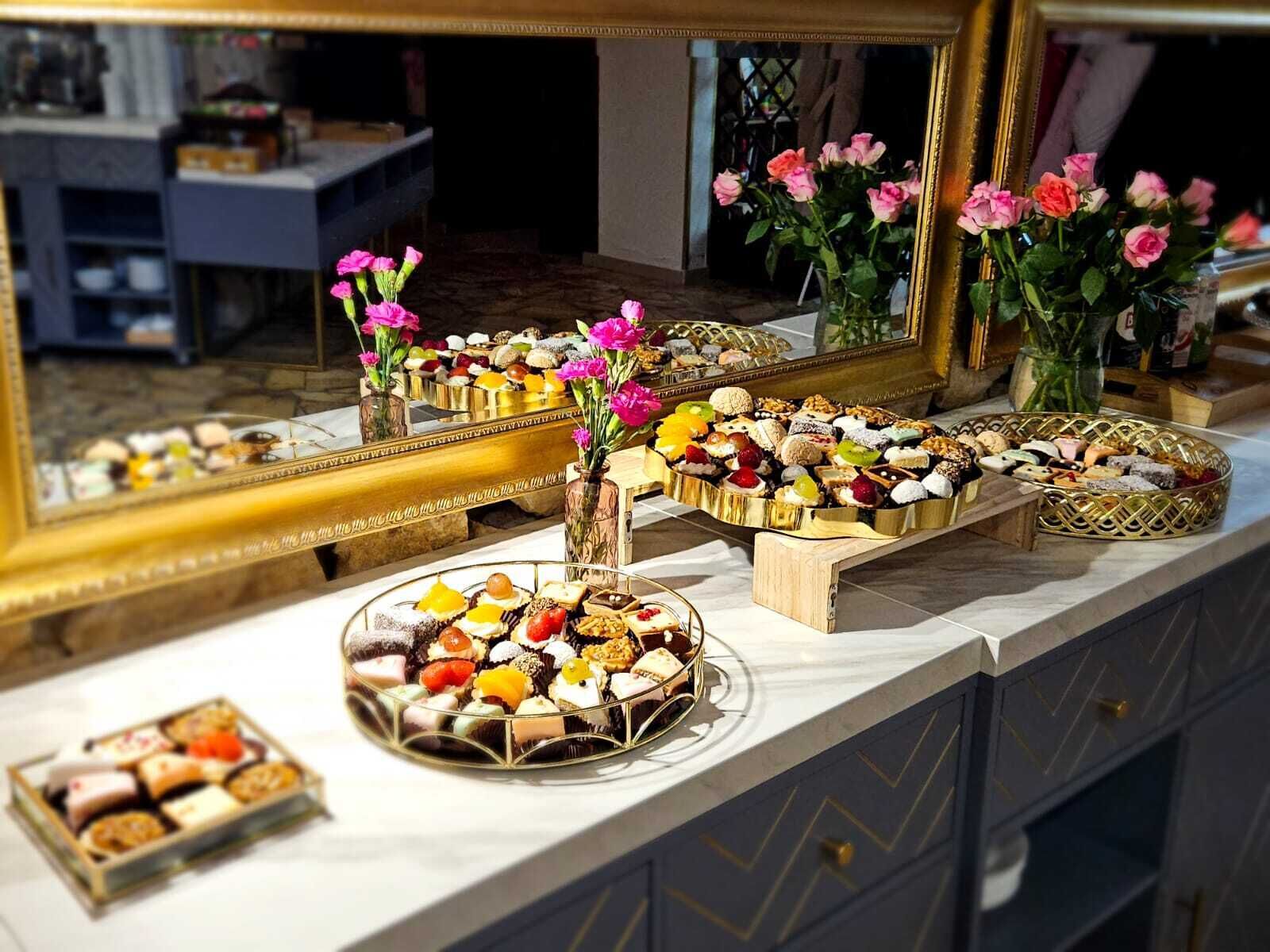 Una mesa de buffet con diversos postres y pasteles, un espejo que refleja la presentación, y flores rosadas en jarrones.
