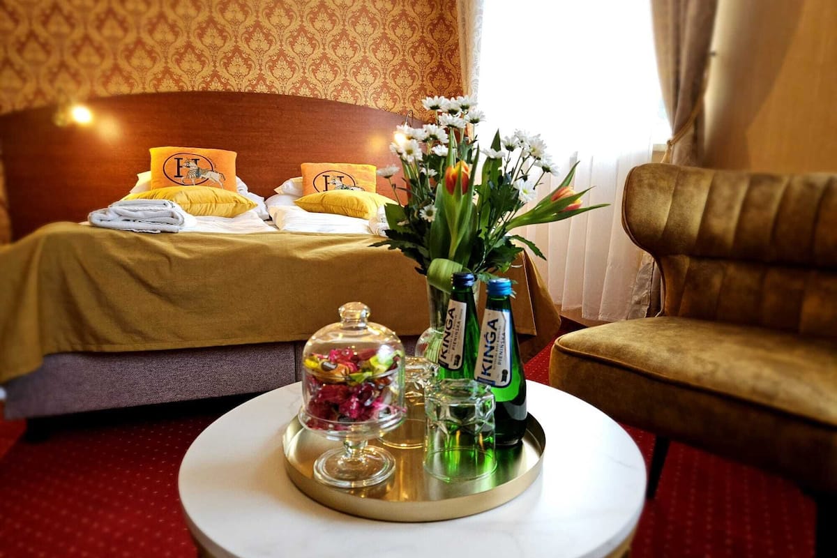 A hotel room with a neatly made bed, a round table with a glass jar of candies, two bottles of KINGA beer, and a vase of flowers.