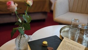 Hotellihuone, jossa on sänky, sohva, pieni pöytä maljakko täynnä kukkia, tarjotinalus laseineen ja ruokalista.