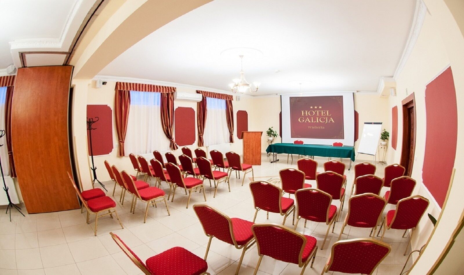Uma sala de conferências com fileiras de cadeiras vermelhas, um púlpito e uma tela exibindo 'HOTEL GALICJA'.